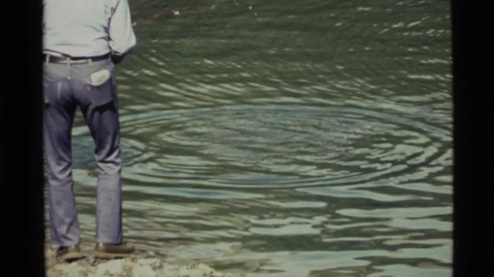 The frame crops the man at the left edge while the lake surface fills the view with widening ripples near the rocky shore at Holland Lake.