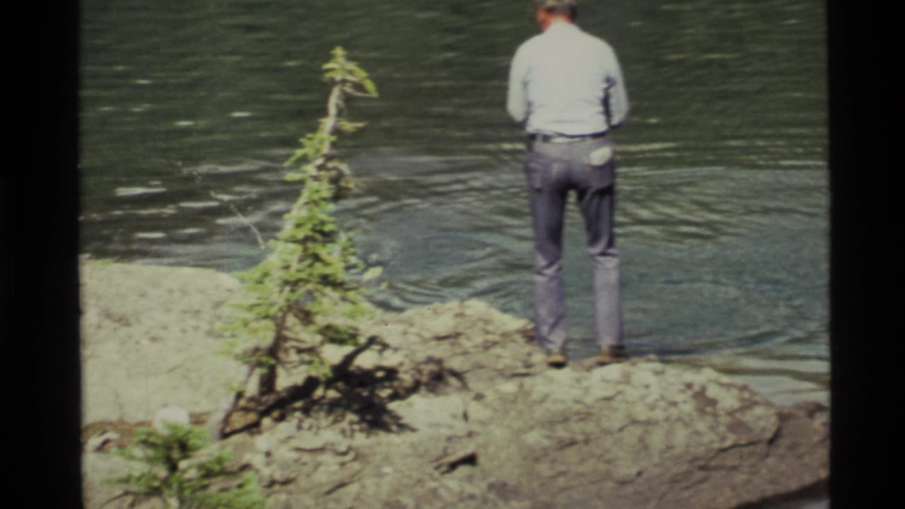 1972 Holland Lake, Montana home-movie frame showing a man from behind fishing on a rocky shore near a small evergreen.