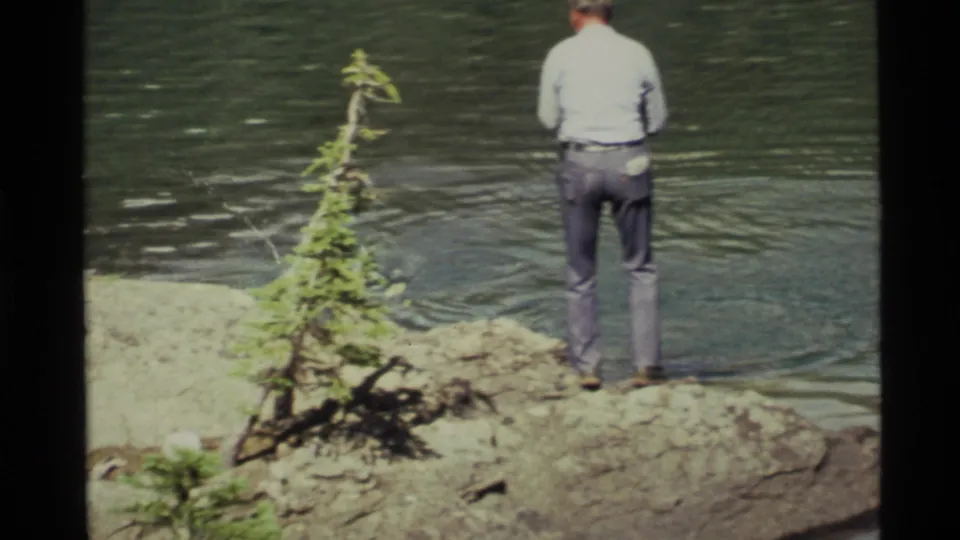 1972 Holland Lake, Montana home-movie frame showing a man from behind fishing on a rocky shore near a small evergreen.