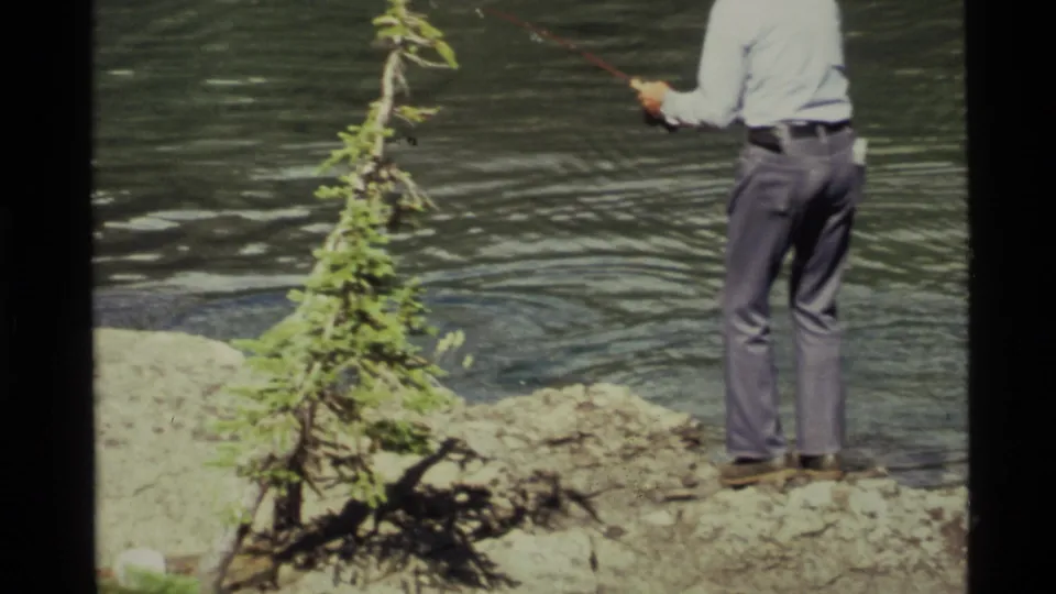 1972 Holland Lake frame with a partly cropped fisherman holding a rod on a rocky shore beside rippling water.