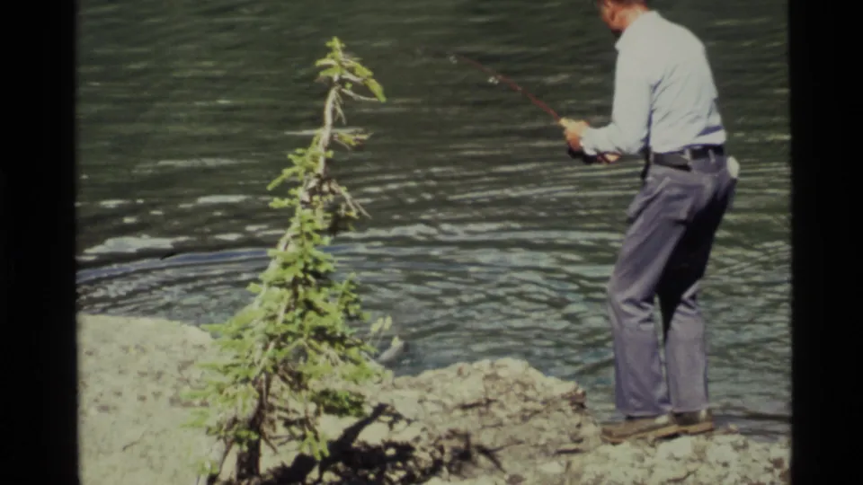 1972 Holland Lake home-movie frame showing a man fishing from a rock bank with a sapling in the foreground.