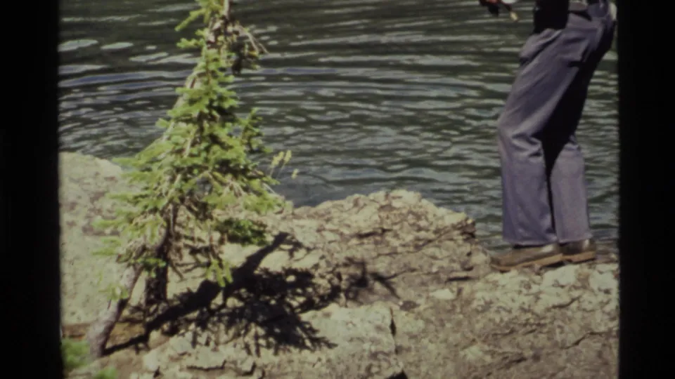 Cropped 1972 Holland Lake frame with the fisherman's legs on a rocky ledge near a small evergreen and water.