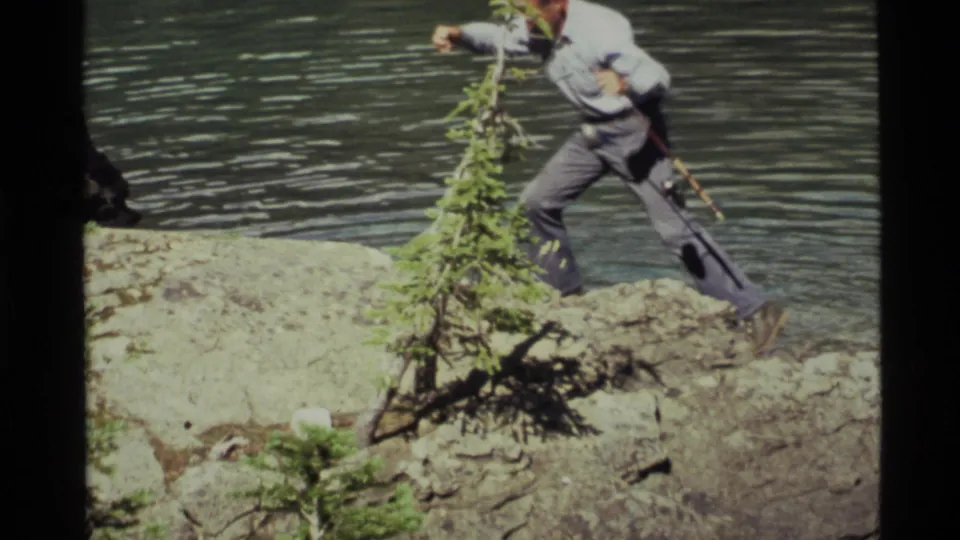 The fisherman leans toward a small evergreen for balance on the rocky Holland Lake shoreline.