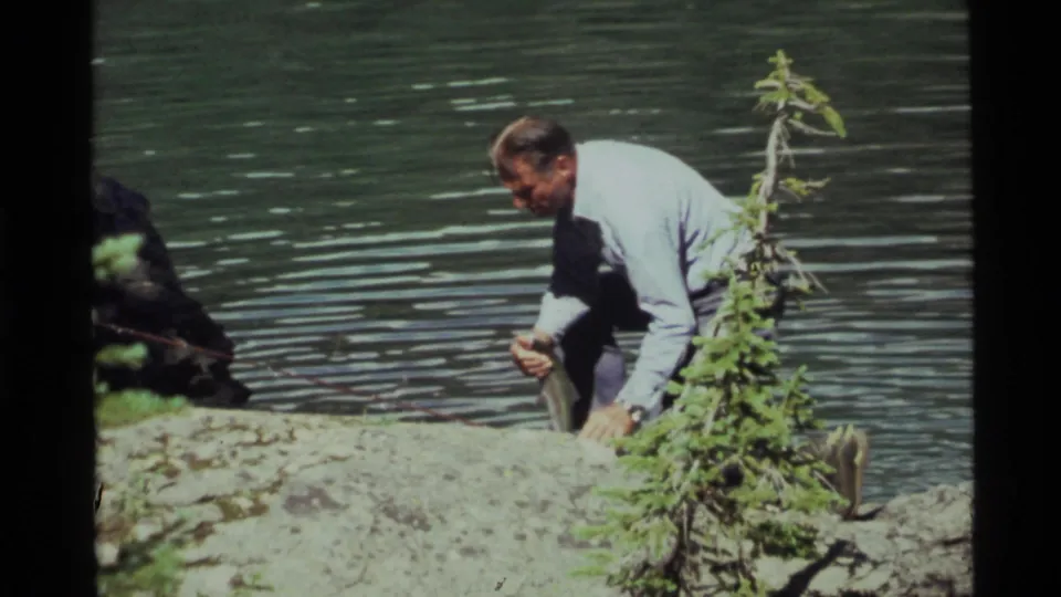 At Holland Lake, Montana in 1972, the man kneels at the shore and holds a small fish with rippled water behind him.