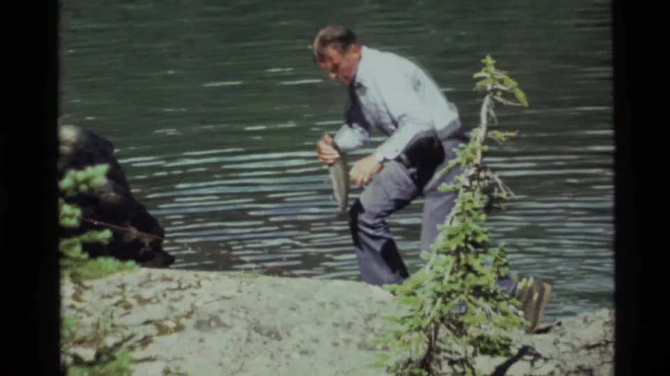 At Holland Lake, Montana in 1972, the man rises from the rocky shore while holding a small fish against the lake background.