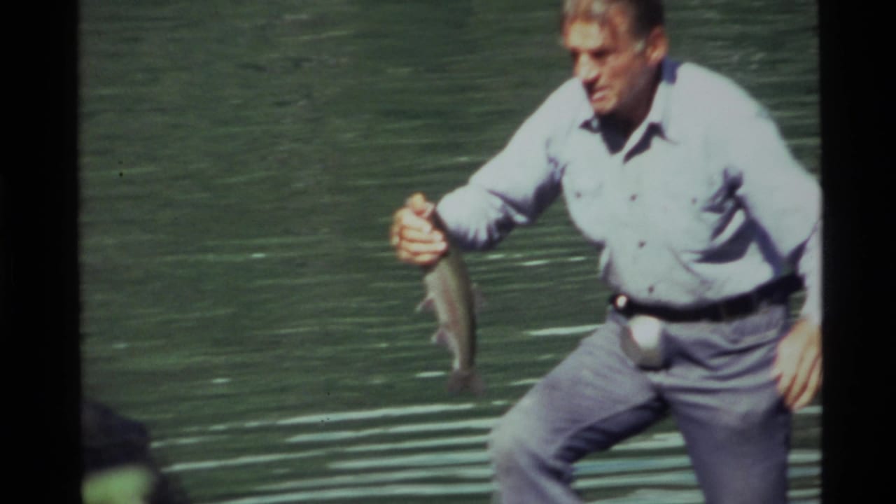 At Holland Lake in 1972, a man in a light shirt crouches at the waterline while holding a freshly caught fish.