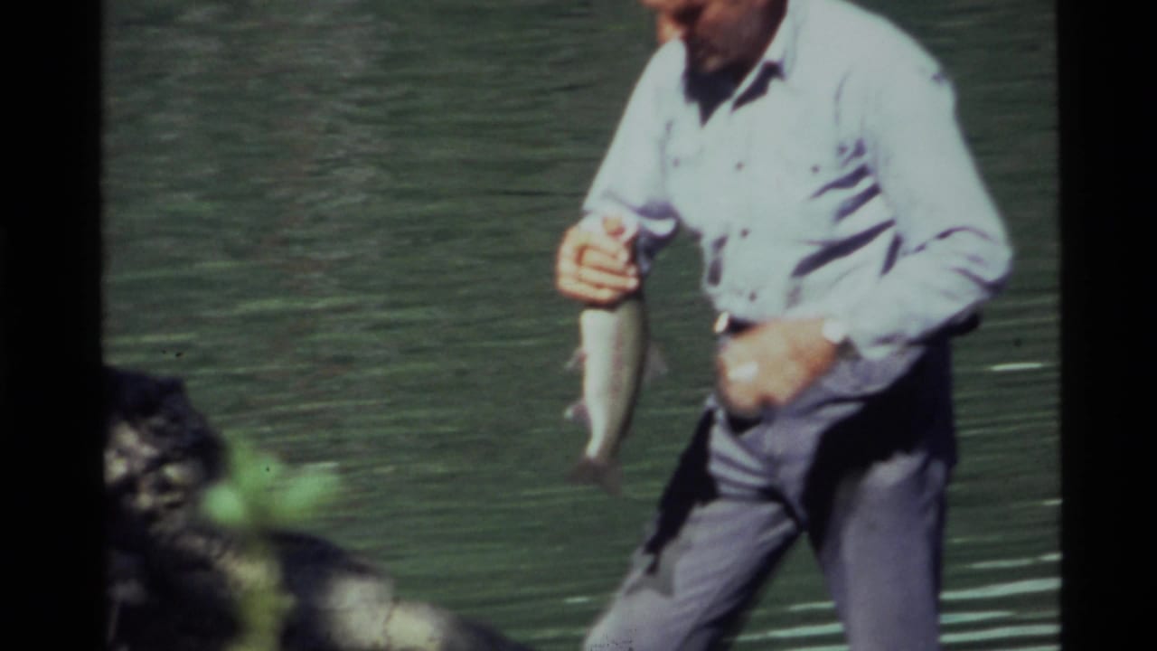 At Holland Lake in 1972, the man stands near the shore and grips the fish with lake water behind him.