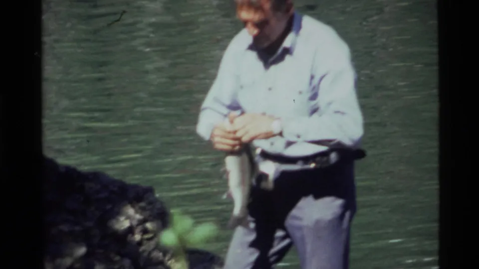 The 1972 Holland Lake footage shows the man looking down as he handles the fish close to the rocky shore.