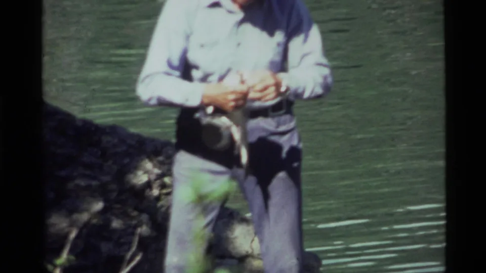 In the 1972 Holland Lake home movie, the man holds the fish with both hands as sunlight washes the lake backdrop.