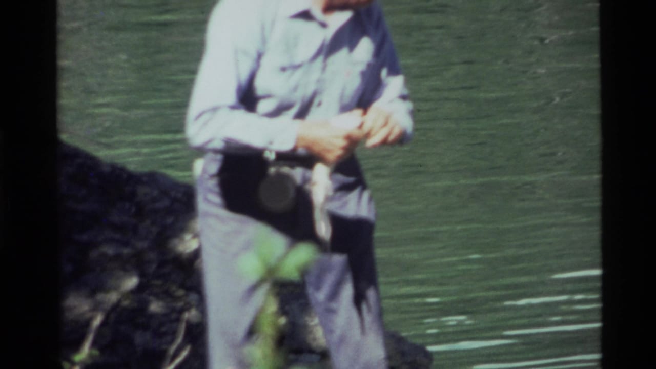 1972 Holland Lake home-movie frame showing a person near green water, partly cropped, with a dark shoreline rock beside them.