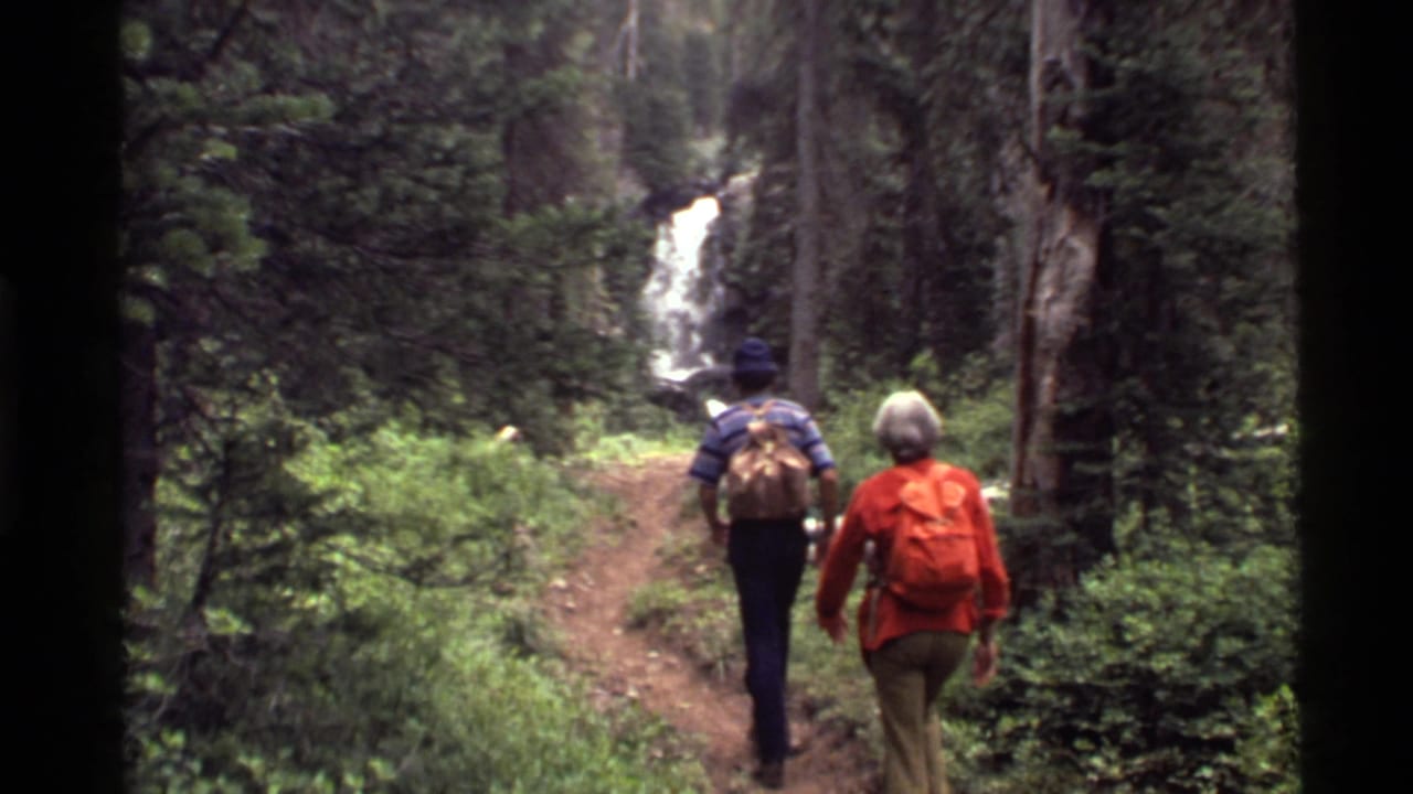 Two backpacked hikers walk on a forest trail toward a waterfall.