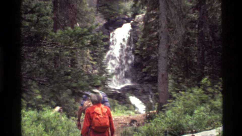 The hikers appear near the waterfall and stream corridor, filmed from behind in the wooded Holland Lake setting.
