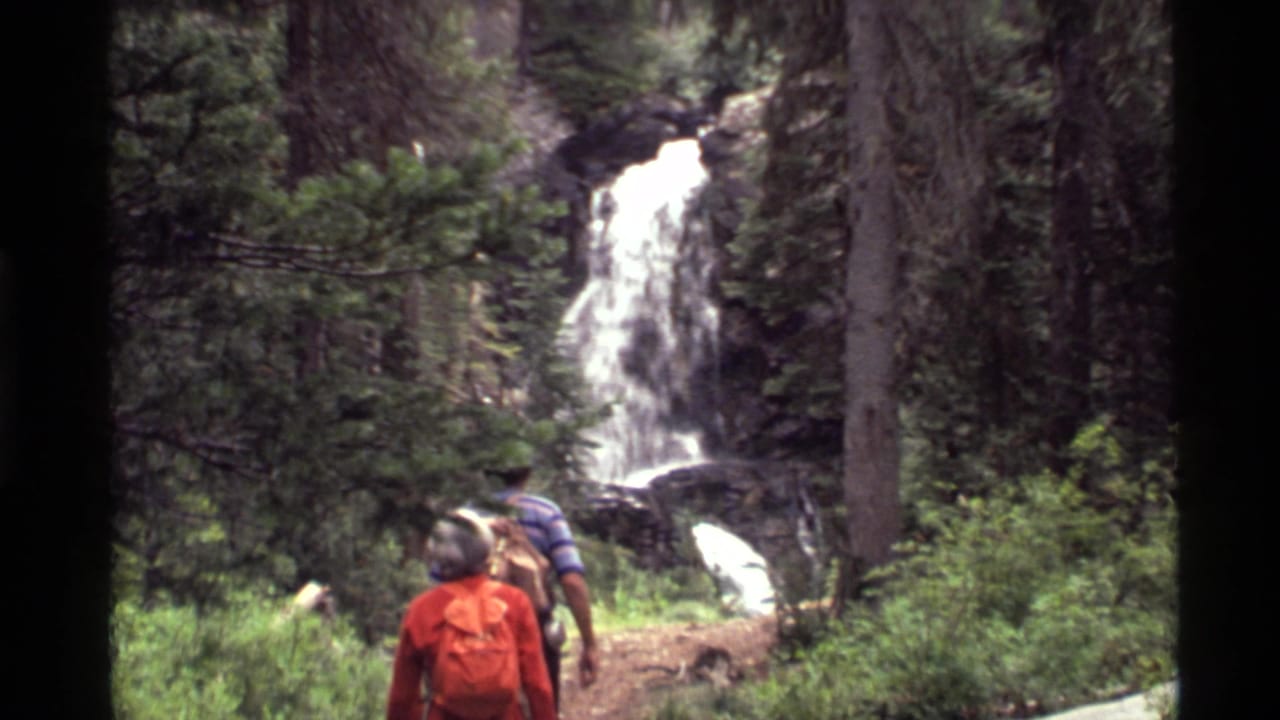 1972 Holland Lake, Montana home-movie frame showing two hikers from behind on a dirt trail below a forest waterfall.