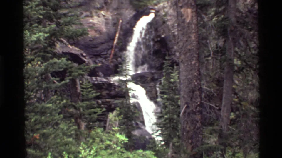 1972 Holland Lake, Montana frame centered on a white waterfall cascading through evergreens and rocky cliffs.