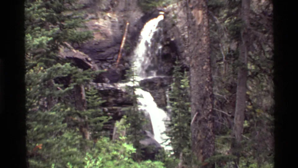 1972 Holland Lake, Montana frame showing the waterfall continuing through dense evergreens, with a large tree trunk at the right edge.