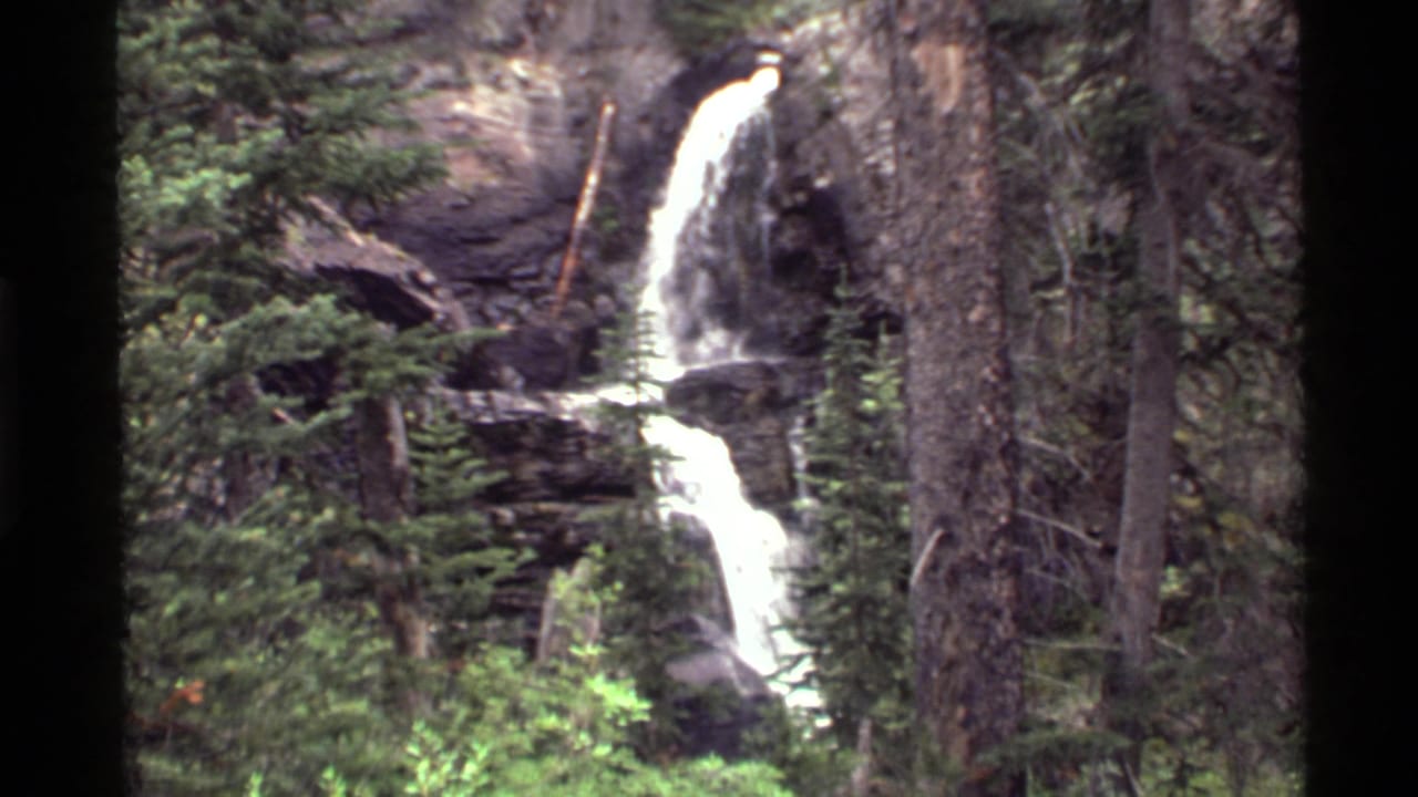 1972 8mm view of a waterfall near Holland Lake, Montana, dropping through dark rock and conifer forest.