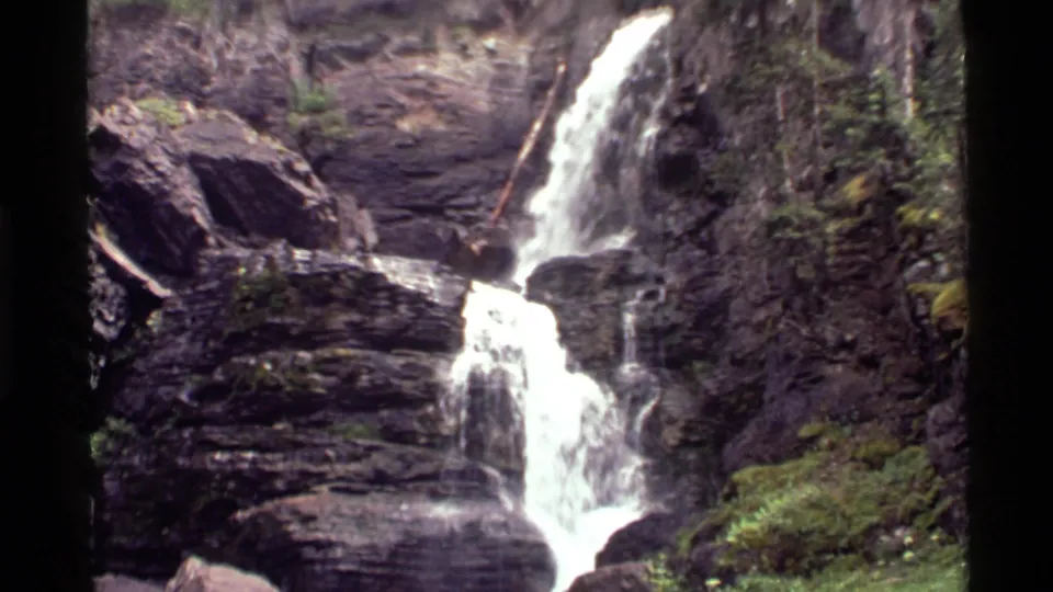 Water pours down a rocky slope near Holland Lake, Montana, with moss, logs, and wet stone visible in the 8mm frame.