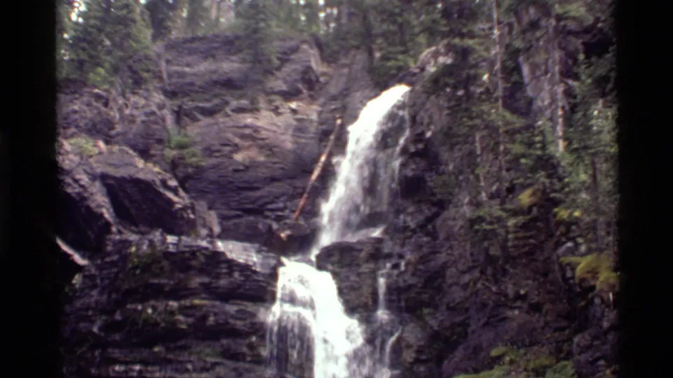 A small Montana mountain waterfall descends through a rocky channel, with moss, trees, and bright water highlights visible.