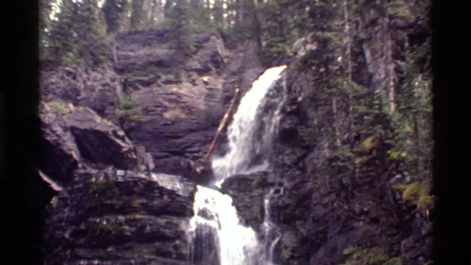 End frame of the Holland Lake waterfall view, showing white water over dark rock ledges and green forest growth nearby.
