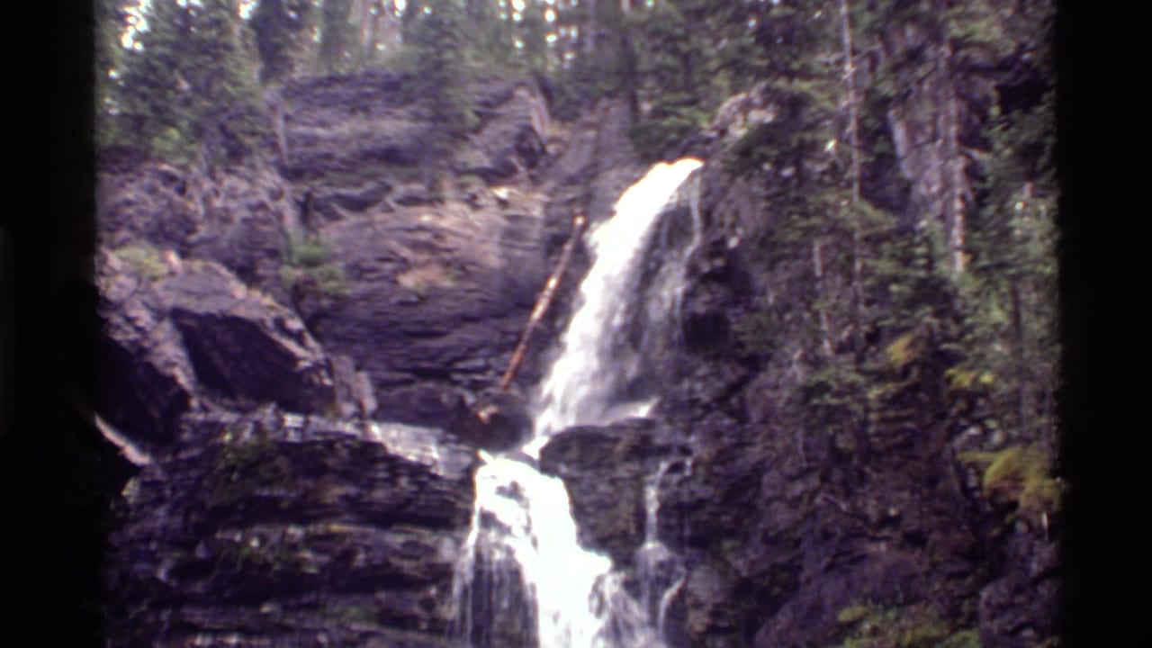 1972 8mm view of a waterfall cascading over dark rock at Holland Lake, Montana, framed by evergreen forest.