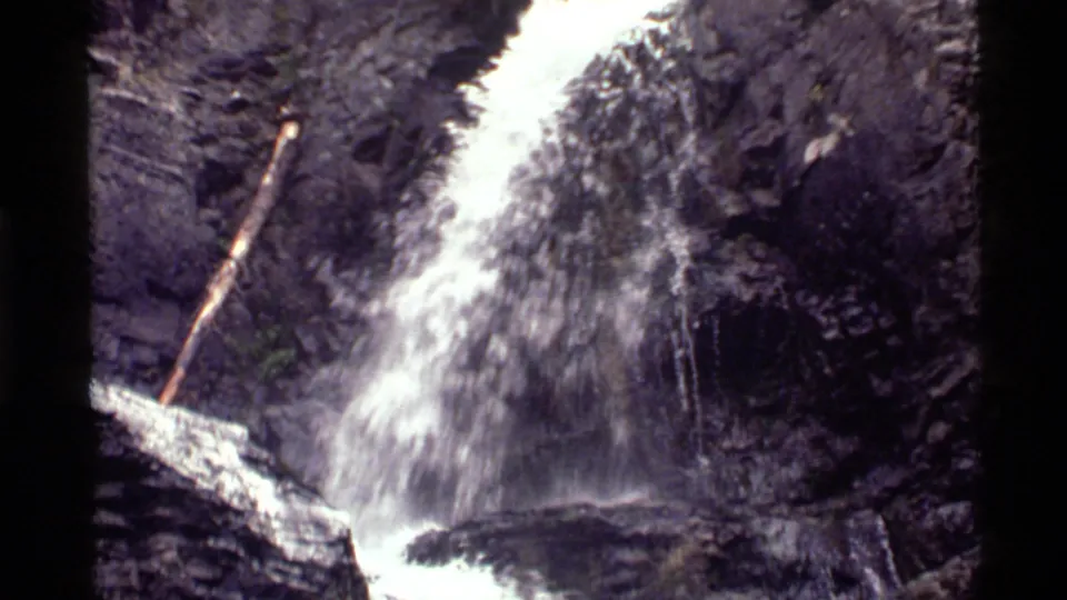 Closer 8mm frame of the Holland Lake waterfall showing water spray, dark stone, and a fallen log near the cascade.