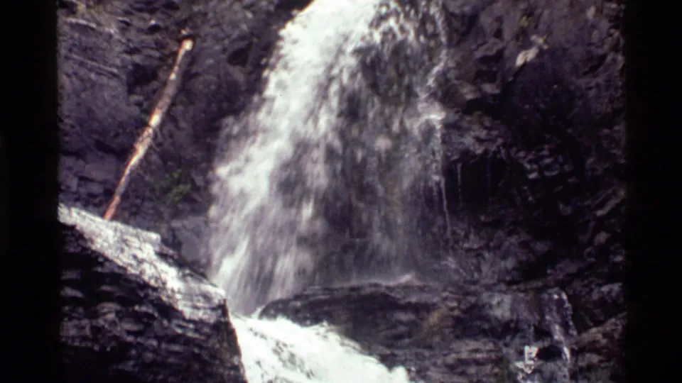 Holland Lake waterfall in 1972 8mm footage, with foaming water, layered rock, and a leaning fallen log.