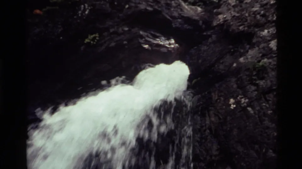 1972 Holland Lake, Montana frame showing water pouring from a rocky mountainside opening into shadowed rock.