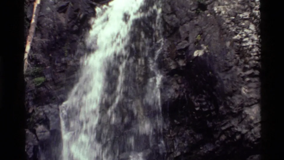 1972 Holland Lake home-movie frame showing a wider view of the waterfall sliding down steep dark rocks with a slender branch nearby.