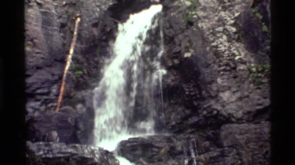 1972 8mm Holland Lake frame with water falling beside a slender branch and breaking against dark ledges at the base of the cascade.