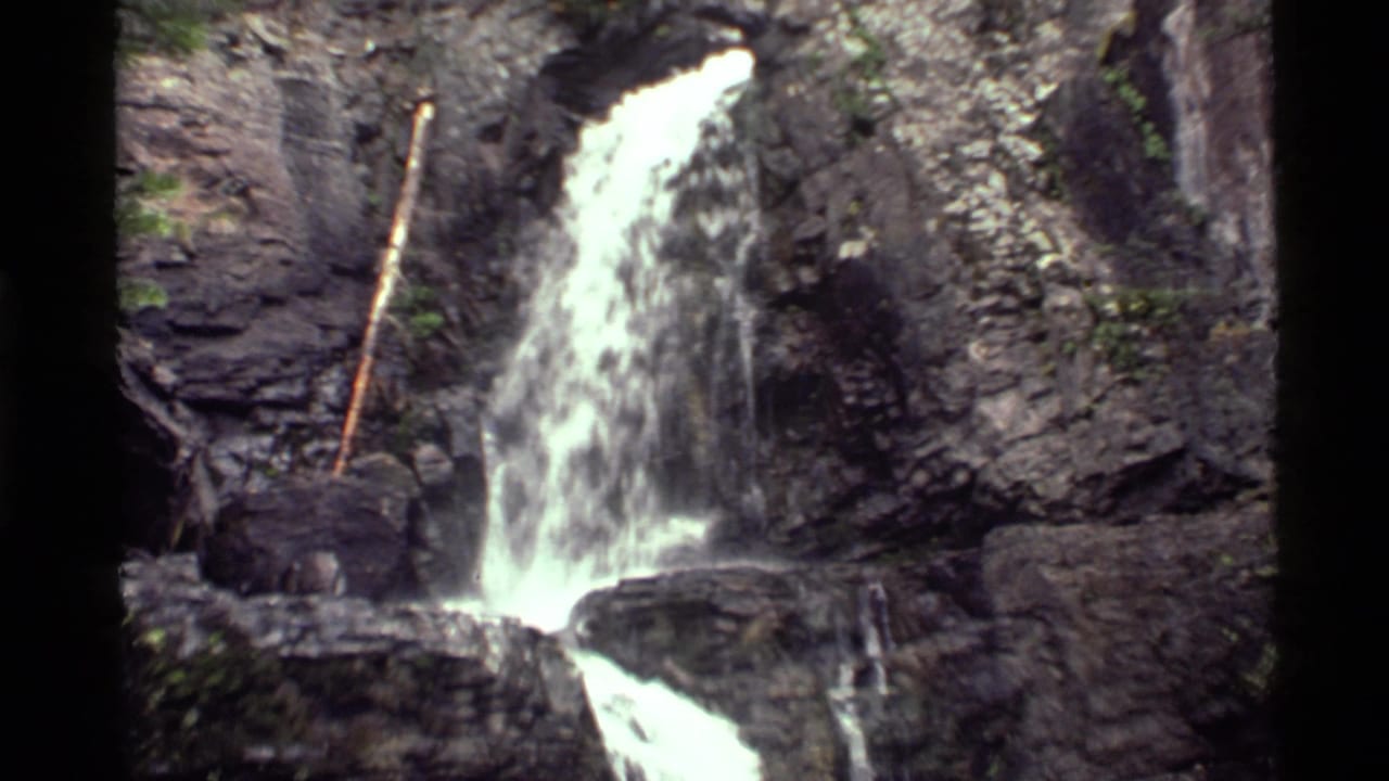 1972 Holland Lake home-movie frame showing a small waterfall dropping over dark layered rock with greenery at the edges.