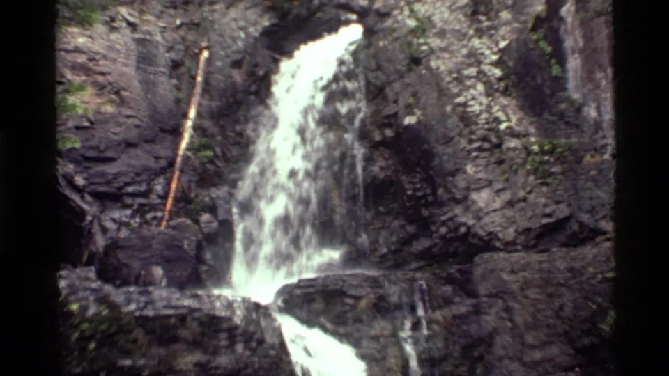 1972 Holland Lake home-movie frame showing a small waterfall dropping over dark layered rock with greenery at the edges.
