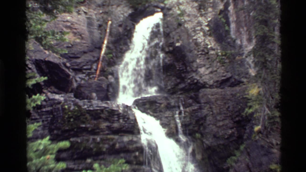Holland Lake, Montana waterfall seen wider, with bright water splitting across dark rock ledges and evergreen branches nearby.