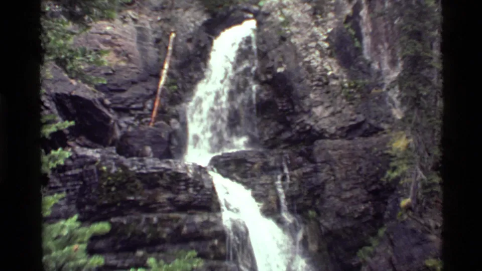 Holland Lake, Montana waterfall seen wider, with bright water splitting across dark rock ledges and evergreen branches nearby.
