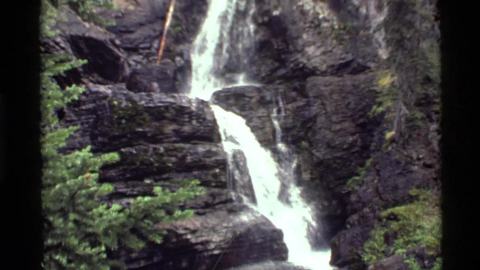 Wider 1972 Holland Lake frame showing the upper and lower waterfall courses among layered rocks and nearby evergreen growth.