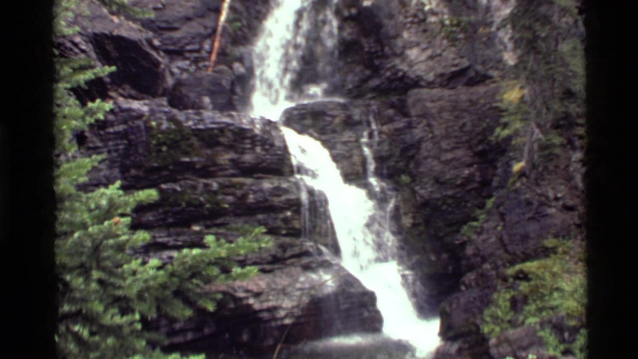 1972 8mm view of a waterfall at Holland Lake, Montana, flowing over dark layered cliffs with conifers and vegetation nearby.