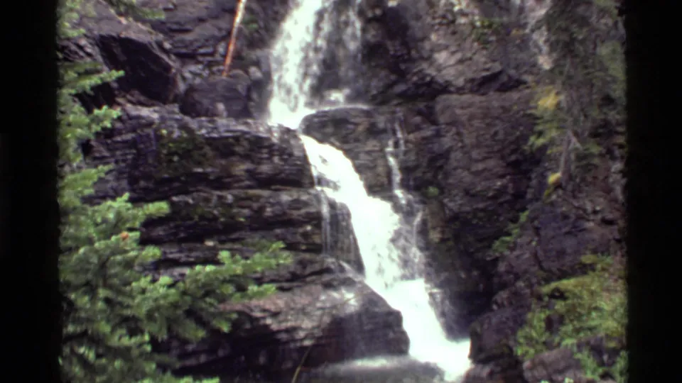 1972 8mm view of a waterfall at Holland Lake, Montana, flowing over dark layered cliffs with conifers and vegetation nearby.