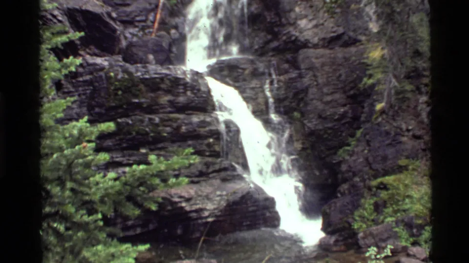 1972 8mm frame from Holland Lake, Montana, showing white water descending through a rocky cliff channel with conifers around it.