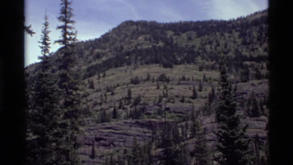 1972 8mm view near Holland Lake, Montana, showing a forested mountain slope with conifers, open rocky terrain, and pale sky.