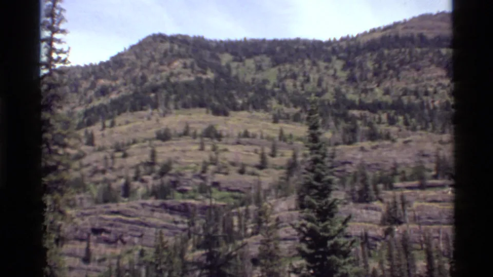 1972 Holland Lake, Montana home-movie frame showing a steep pine-dotted slope with rocky bands and dark film borders.