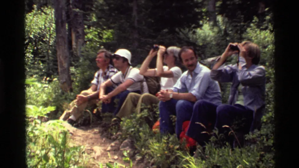 At Holland Lake, Montana in 1972, adults sit together on a trail while several use binoculars in the forest.