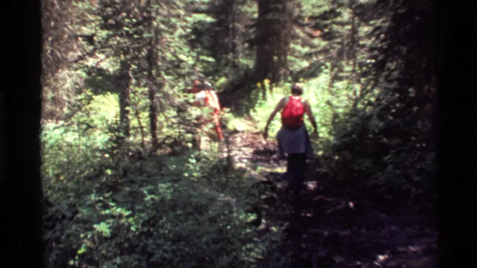 Two hikers continue along the Holland Lake woodland trail, seen from behind as one carries a bright red backpack.
