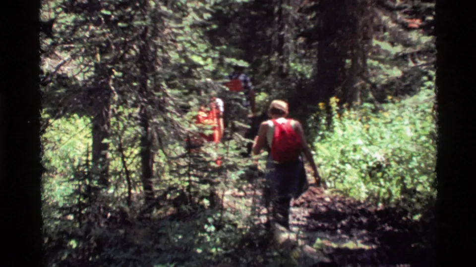 Holland Lake hiking scene with a red-backpack figure following companions through dense 1970s forest footage.