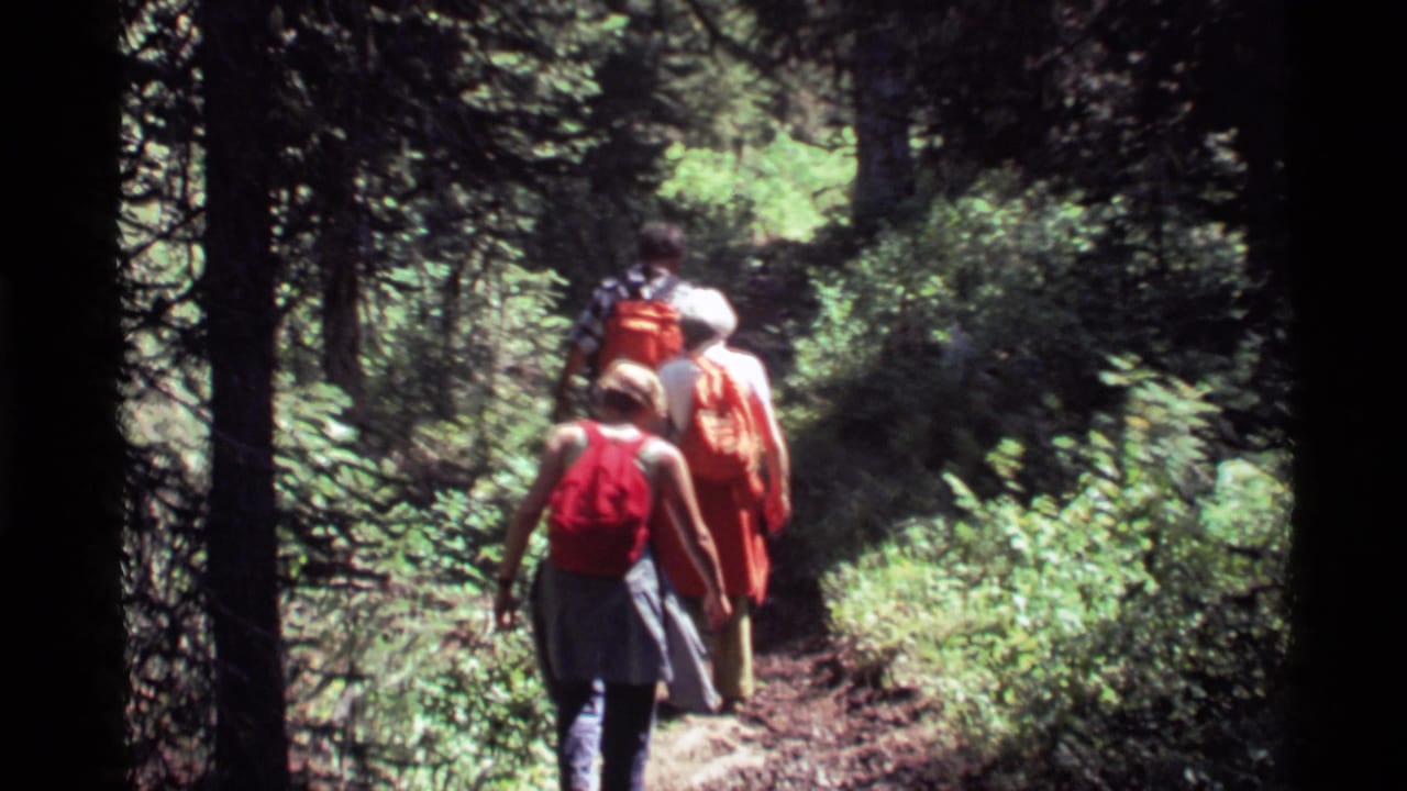 Three people hike away from the camera on a narrow forest trail at Holland Lake, Montana, in 1972 home-movie footage.