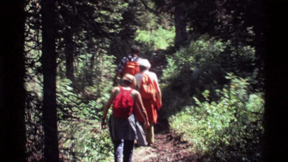 Three people hike away from the camera on a narrow forest trail at Holland Lake, Montana, in 1972 home-movie footage.