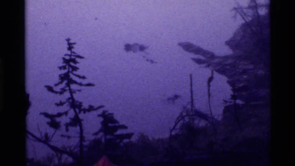 Visitor-shot 8mm frame from Holland Lake, Montana, with a dark evergreen, brush, and foggy purple-blue shoreline atmosphere.
