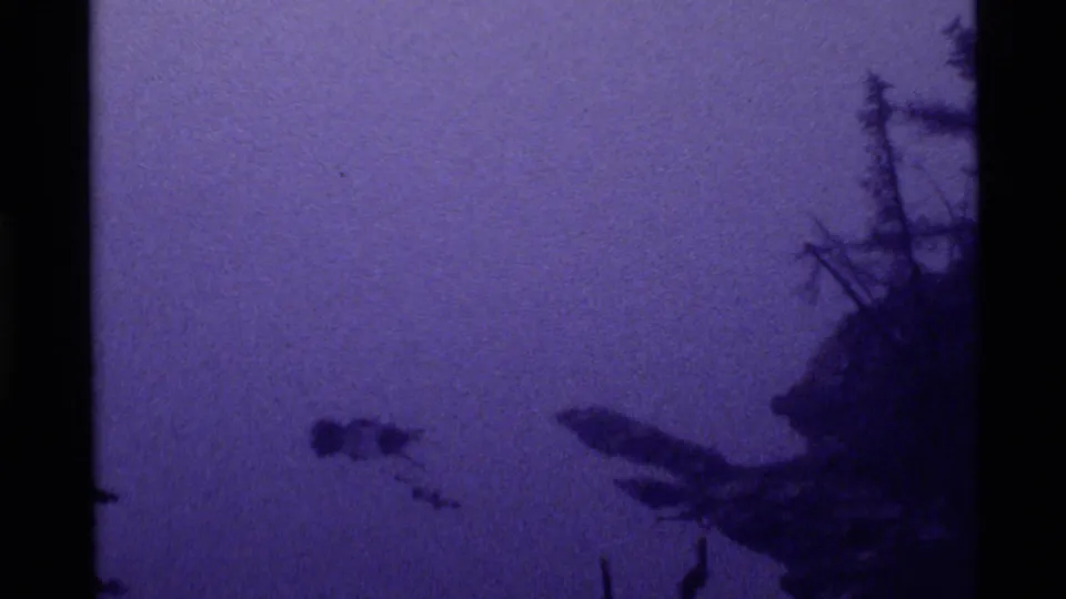 1972 Holland Lake 8mm still with a purple color cast, dark lakeshore trees, and a small floating form on the quiet water.