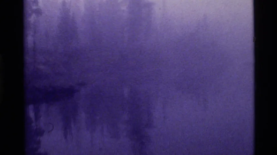 1972 Holland Lake home-movie frame showing mist, soft reflections, and dark evergreen forms along the edge of the water.