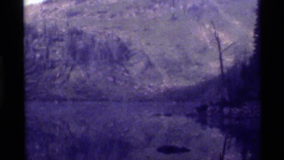 Soft 1972 8mm frame of Holland Lake with a dark shape on the water, shadowed evergreens, bare tree trunks, and a snowy mountain rising behind.