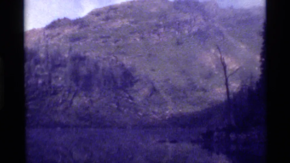 Final 8mm frame looks across dim Holland Lake toward a tree-lined shore and snow-dusted mountain, with strong purple-blue fading and soft exposure.