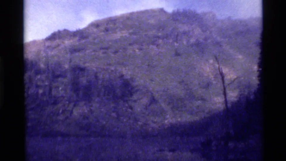 1972 Holland Lake, Montana footage showing a rocky, tree-covered mountain slope under a pale sky with soft 8mm grain.